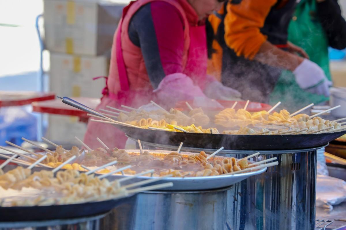 Street vendor preparing skewers in large pots, steam rising from the food