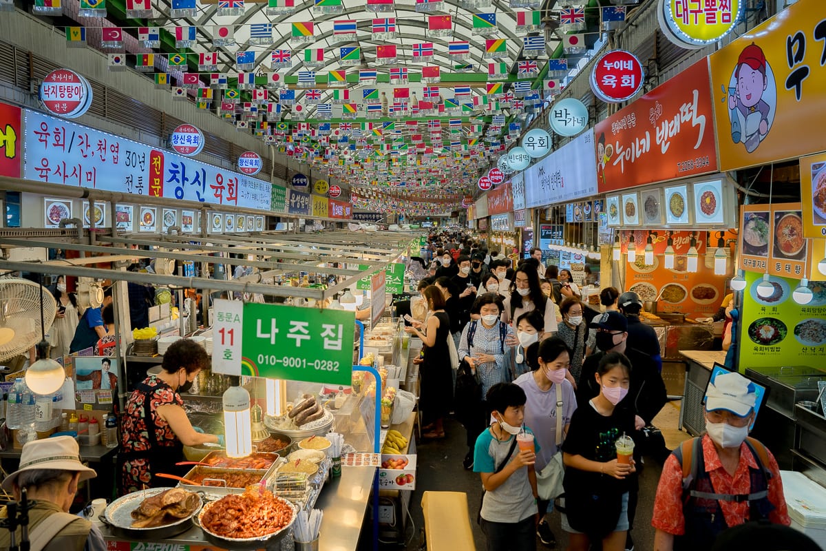 Vibrant indoor market with masked crowd, food stalls, and colorful flags overhead.
